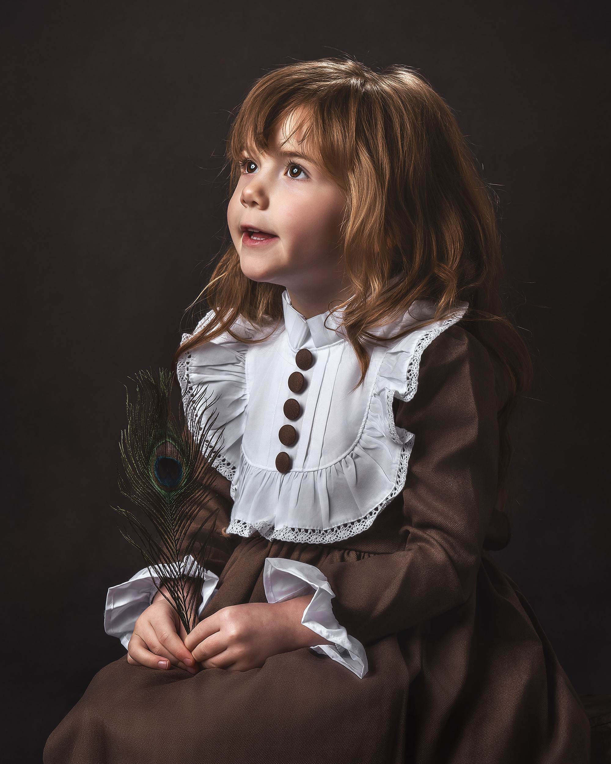 A fine art child portrait by AMM Fine Art Studio, featuring a young girl in a brown Victorian-style heirloom dress holding a peacock feather. This commissioned study utilizes painterly studio lighting to create a timeless masterpiece for luxury clients in Medina, Bellevue, and Kirkland.